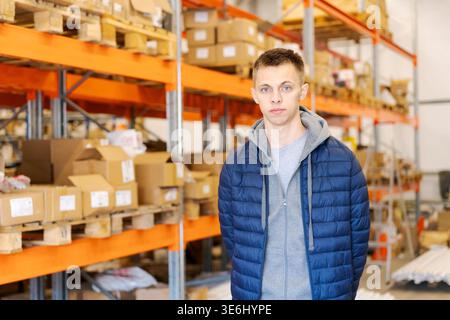 A focused young man standing in a large distribution warehouse with orange racks, wooden pallets, and stock boxes. Concept of logistics, storage, and e-commerce inventory. Stock Photo