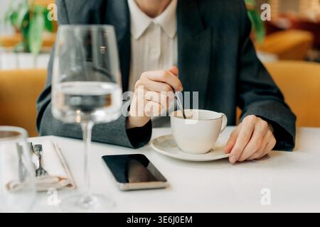 Close-up of an unrecognizable woman's hands placing sugar in coffee in a restaurant while her mobile phone lies nearby on the table. Stock Photo