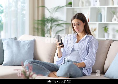 Disappointed woman in blue checking phone sitting on a couch at home Stock Photo
