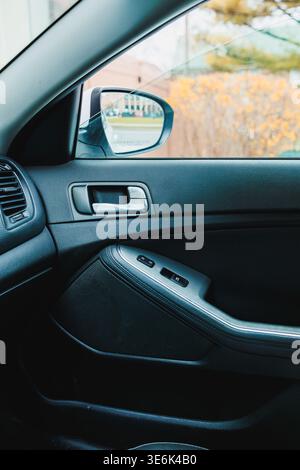 A person's view inside a car shows the door with window controls and a side mirror. The car is parked, and trees are blurred in the background. Stock Photo
