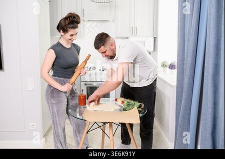 Happy Couple Cooking Homemade Pizza Together in Bright Kitchen, Man Rolling Dough and Woman Holding Rolling Pin, Fun Domestic Life and Teamwork Concep Stock Photo