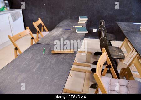 Wooden desks with scattered books and pencils arranged in rows, soft natural lighting, black chalkboard wall background, empty chairs with cushions, q Stock Photo