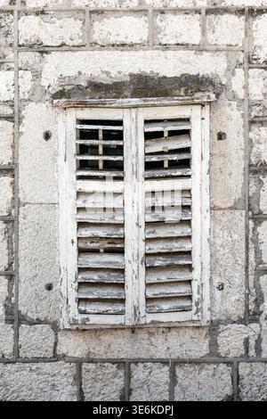 Weathered white wooden shutters set in an old stone wall convey rustic charm and aged character. Peeling paint, cracks and rough masonry create a vint Stock Photo