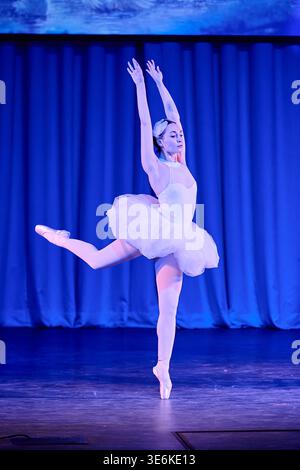Elegant ballerina in white tutu and pointe shoes executing arabesque pose, blue stage lighting, dark curtain backdrop, classical ballet performance at Stock Photo