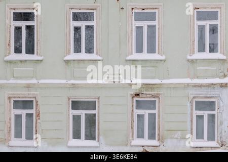 Snow lines the sills of eight white-framed windows set in a pale green, weathered facade, creating a quiet, cold winter atmosphere with architectural Stock Photo