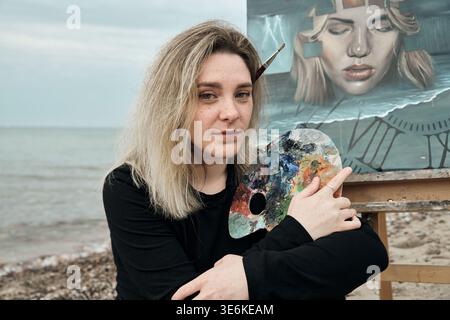 Young adult female artist with blonde hair holds colorful palette and brush near easel showing surreal portrait while standing on Rocky Beach under ov Stock Photo