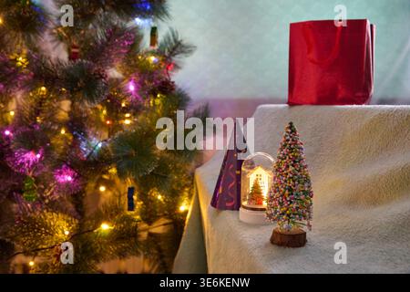 Colorful christmas tree lights, decorated miniature christmas tree, snow globe, party hat, and shiny red gift bag on beige draped table in warm cozy r Stock Photo