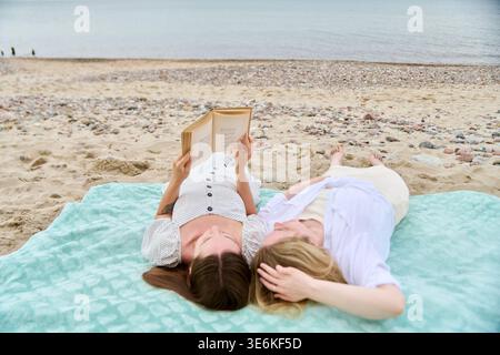 Two young women lying on turquoise blanket on sandy beach with pebbles, one reading book while other rests head on her hand, calm sea in background Stock Photo