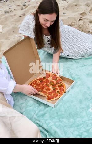 Young woman in white dress enjoys relaxing beach picnic while sharing pepperoni and jalapeno pizza inside open cardboard box on soft pastel green blan Stock Photo