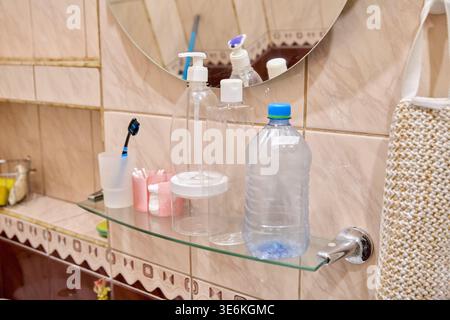 Clear plastic pump bottles and transparent water bottle arranged on glass shelf in beige tiled bathroom with round mirror and black toothbrush in cup Stock Photo