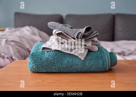 Plush turquoise towel with neatly folded smaller gray and beige towels stacked on top, placed on warm wooden surface in living room setting with muted Stock Photo