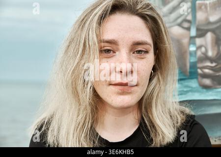 Adult woman painter with light blonde hair and neutral expression stands outdoors near painted mural. Soft lighting highlights natural features and fr Stock Photo