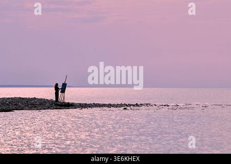 Adult male artist with dark hair paints on easel set on rocky shoreline under soft pink evening sky. Peaceful coastal scene with tranquil water and di Stock Photo