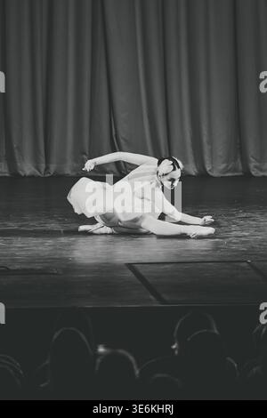 Poised ballerina in white tutu extends leg and arm on dark wooden floor with heavy curtain backdrop, conveying delicate emotion amid theatrical spotli Stock Photo