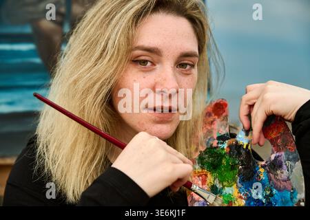 Adult female artist with blonde hair and freckles holds paintbrush and vibrant palette, working on canvas in well-lit studio. Focused expression captu Stock Photo