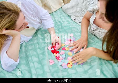 Two adult women engage in sorting multicolored paper hearts while sitting on soft teal blanket, reflecting themes of female friendship and love in coz Stock Photo