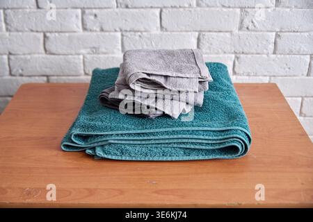 Soft folded gray washcloths neatly stacked on thick teal bath towel placed on smooth wooden table against textured white brick wall background Stock Photo