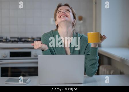 Smiling woman looking up with clenched fists expressing excitement, anticipation or gratitude sits at home with laptop. Happy young female joyful in g Stock Photo