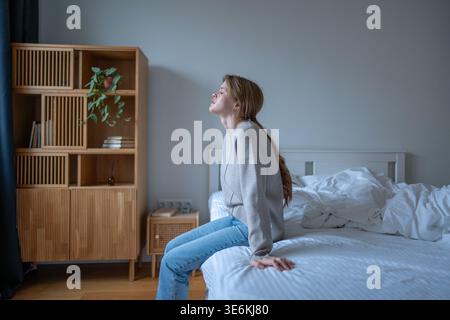 Drained depressed young woman sits on unmade bed wearing outdoor clothes feeling burnout, mental exhaustion and nervous breakdown after stressful day. Stock Photo