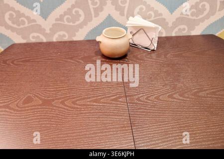 Smooth reddish-brown wood grain table with small cream ceramic pot and silver napkin holder containing white napkins, set against beige and blue patte Stock Photo