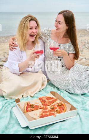 Two young women share laughter while sitting on blanket at Rocky Beach, drinking pink wine and eating pepperoni pizza, capturing joyful and relaxed mo Stock Photo