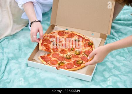 Close-up of two young adult women hands taking slices from pepperoni pizza with jalapenos on picnic blanket outdoors in natural daylight. Casual femal Stock Photo