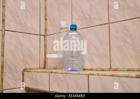Clear plastic water bottle with blue cap standing on brown patterned tiled bathroom shelf next to translucent white containers, soft natural lighting Stock Photo