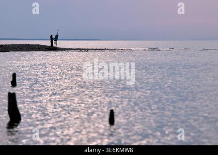 Adult man paints on easel at lake shore under pastel dusk sky, capturing tranquil waters and serene landscape in peaceful solitude Stock Photo