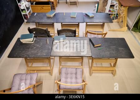 Dark wooden desks with scattered books and pens arranged in two rows on beige tiled floor, cushioned chairs facing desks, neutral classroom environmen Stock Photo