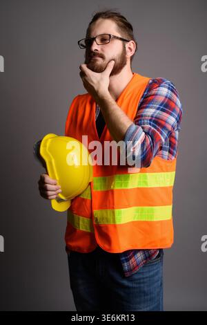 Studio portrait of a pensive bearded man in an orange safety vest holding a yellow hard hat and touching his chin. Ideal for construction planning, vi Stock Photo