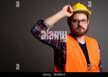 Studio portrait of a bearded Caucasian man in an orange reflective safety vest adjusting his yellow hard hat. Ideal for industrial labor, engineering, Stock Photo