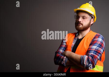 Studio portrait of a serious bearded man in an orange safety vest and yellow hard hat looking down with arms folded. Features large copy space for eng Stock Photo