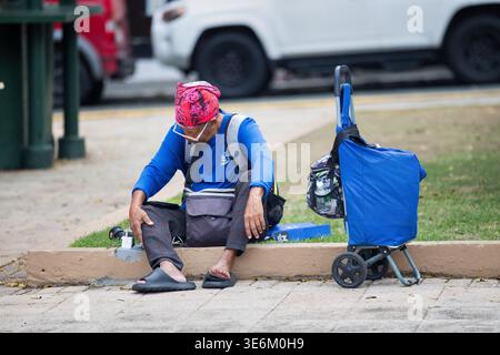 Homeless man  with blue jacket and red hat sitting on pavement in the Caribbean with his trolley Stock Photo