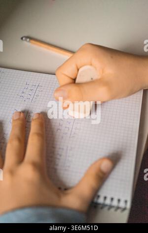 Close-up of a child's hands using an eraser to erase math operations written in a graph paper notebook. Concept of studying, learning, and basic educa Stock Photo