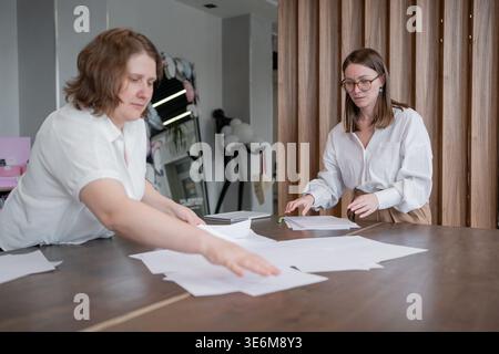 Two women reviewing and organizing documents together at tabletop in light open office. Reviewing, organizing, documents, tabletop, women, office. Stock Photo