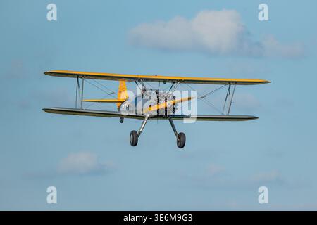 A vintage yellow biplane in flight against a clear blue sky during an outdoor airshow display. Open cockpit, radial engine and classic struts convey n Stock Photo