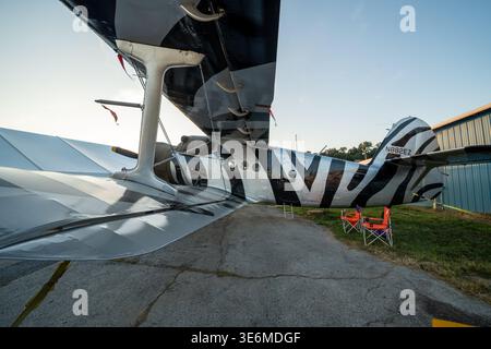 Zebra-striped amphibious airplane parked beside a hangar at a small airport, photographed from beneath the wing in warm late light. Details include ta Stock Photo