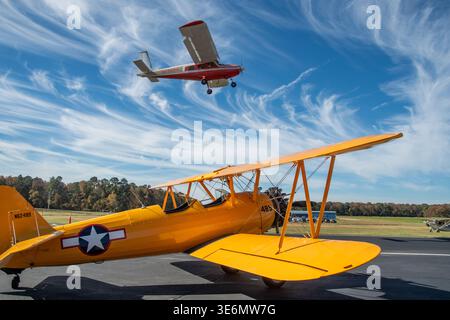Yellow vintage biplane parked on an airfield ramp while a red small Cessna flies overhead against wispy blue sky. Autumn treeline and hangars visible, Stock Photo