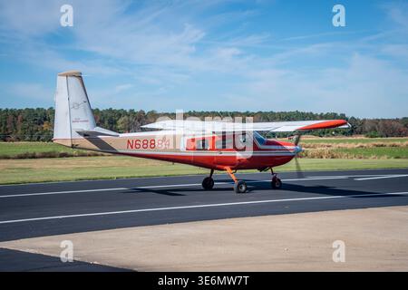 Red-and-white single-engine small airplane taxis on a runway at a rural airport beneath a clear blue sky, with grassy fields and a distant tree line c Stock Photo