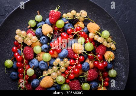 Bright mixed berries in grey bowl, top view on dark table. Organic blueberries, currants, raspberries, gooseberries and mulberries Stock Photo