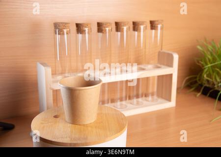 Test tubes are arranged neatly in a holder on a wooden table. A small paper cup is placed on a round wooden surface. Green plants are visible in the background, adding freshness. Stock Photo