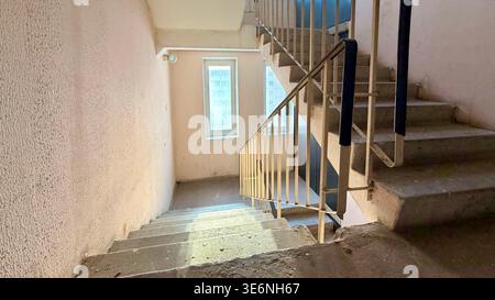 Apartment building stairwell with concrete stairs going down, metal railings and sunlight through window. Stock Photo