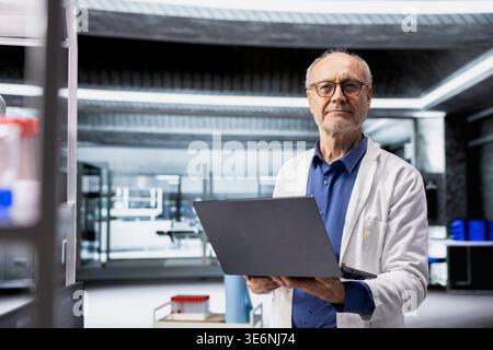 Portrait of senior experienced scientist in white coat holds a laptop to check microbiology experiment data results, working in a professional biolab. Old man chemist working in healthcare. Stock Photo