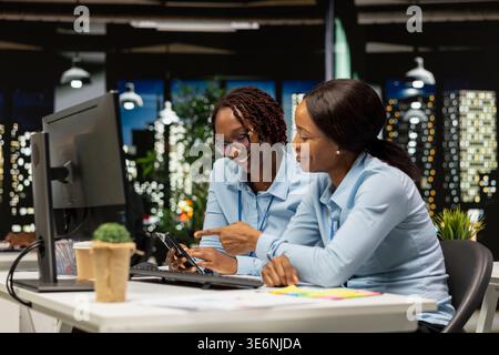 Black women working together with data insights on desktop in modern workspace, developing business solutions with strategy planning and performance metrics analysis for new goals achievement. Stock Photo