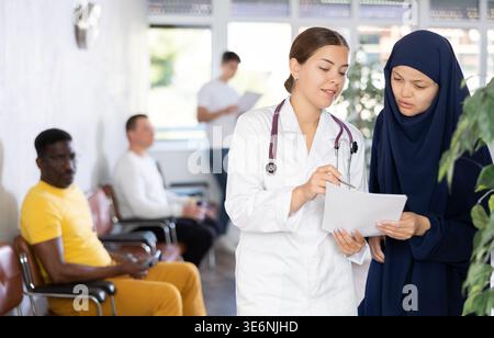 Young female doctor showing papers to Muslim woman in lobby of clinic Stock Photo