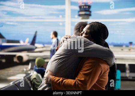 Romantic married couple hugging in busy airport waiting area, creating image of gratitude near tourists and suitcases. Quiet moment before boarding plane for long-distance flight. Stock Photo