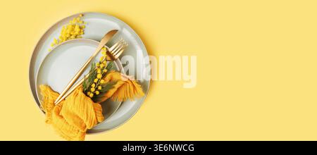 Spring table setting with mimosa flowers and golden cutlery on pastel yellow background Stock Photo