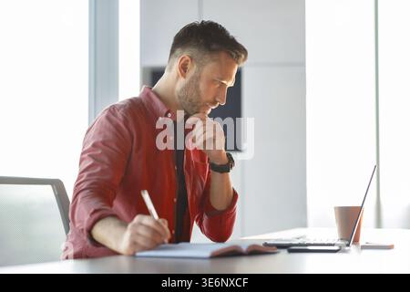 Man Writing Notes While Looking at Computer in a Bright Office Setting in the Afternoon Stock Photo
