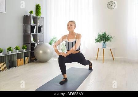 Sporty fit woman performing lunge stretch on mat, stretching body, engaging in exercise Stock Photo