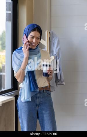 Woman standing by window wearing blue hijab and jeans talking on pink smartphone holding coffee cup Stock Photo